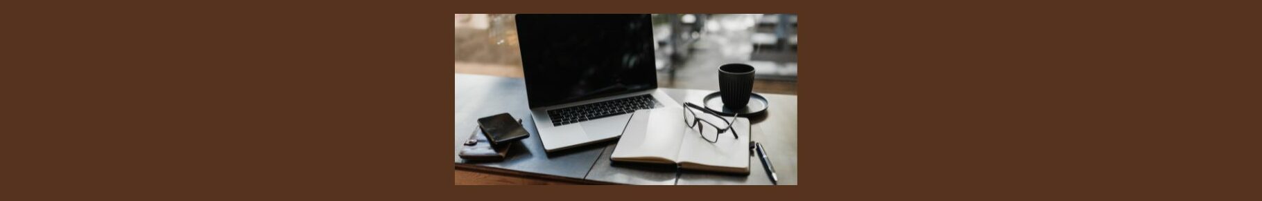 How Does Fiverr Work A Quick Guide. Medium brown background.  a wood table, light gray mat, open laptop, open notebook, white pages, pair of glasses laying on the right side, a black pen beside the notebook, a black saucer and cup above the notebook. 