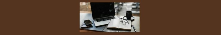 How Does Fiverr Work A Quick Guide. Medium brown background. a wood table, light gray mat, open laptop, open notebook, white pages, pair of glasses laying on the right side, a black pen beside the notebook, a black saucer and cup above the notebook.